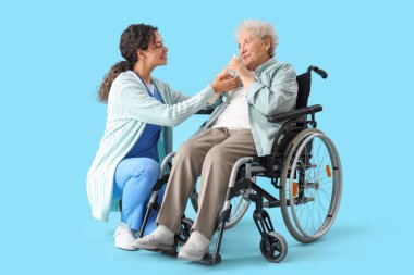 Genç Afro-Amerikalı kadın doktor. Tekerlekli sandalyedeki yaşlı kadına mavi arka planda su veriyor.