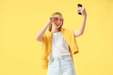 Güneş gözlüklü genç bir kadın sarı arka planda cep telefonuyla selfie çekiyor.