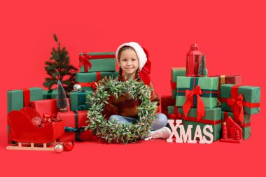 Noel şapkalı, Noel çelengi takan ve kırmızı arka planda oturan tatlı küçük kız.