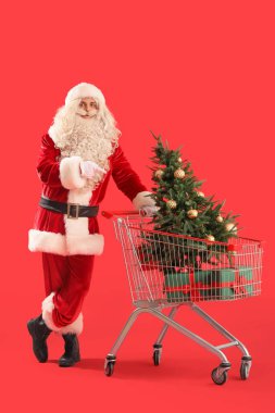 Noel ağacıyla Noel Baba ve kırmızı arka planda alışveriş arabasında hediye kutuları.