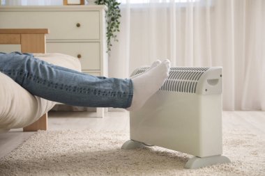 Oturma odasındaki elektrikli ısıtıcının yanındaki kanepede oturan genç adam.