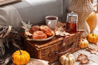 Çaydanlıklı sonbahar kompozisyonu, bir fincan sıcak içecek, lezzetli çörekler ve sepetli balkabakları oturma odasında.