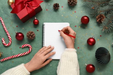 Noel süslemeleri ve hediye kutusu olan bir kadın 2025 yılını grunge yeşil arka planda deftere yazmayı planlıyor.