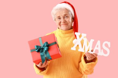 Noel Baba şapkalı yaşlı kadın, pembe arka planda hediye ve kelime XMAS 'ı var.