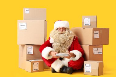 Noel Baba yerde paketler ve sarı arka planda mektuplarla oturuyor.