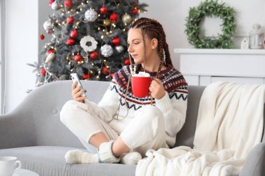 Noel arifesinde evinde bir fincan kakao ve cep telefonu olan genç bir kadın.