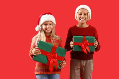 Kırmızı arka planda Noel hediyeleri olan Noel Baba şapkalı çocuklar.