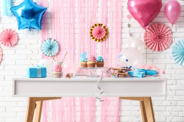 Delicious cupcakes with baby pacifiers and decorations on table against white brick background. Gender reveal party concept clipart