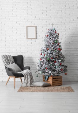 Beyaz tuğla duvara karşı hediyeler ve koltukla süslenmiş Noel ağacı.
