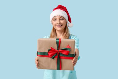 Noel Baba şapkalı olgun kadın doktor mavi arka planda Noel hediyesi kutusu var.