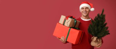Noel Baba şapkalı mutlu genç adam elinde Noel ağacı ve kırmızı arka planda hediye kutuları ve mesaj için yer var.