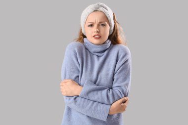 Donmuş, kalın elbiseli, gri arka planda kendine sarılan güzel bir kadın.