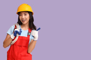 Kablolu genç bayan elektrikçi mor arka planda baş parmağını gösteriyor.