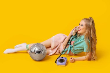 Sarı arka planda retro telefonu ve disko topu olan güzel, genç bir kadın.
