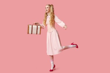 Güzel, genç ve mutlu bir kadın. Pembe arka planda bavulu olan oyuncak bebek gibi giyinmiş.