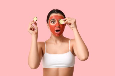 Yüzünde maske ve pembe arka planda salatalık dilimleri olan şaşırmış genç bir kadın.