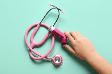 Astım spreyi ve turkuaz arka planda steteskop olan bir çocuk eli.