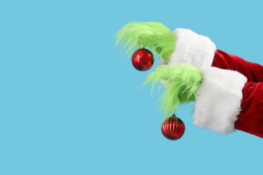 Green hairy hands of creature in Santa costume with red Christmas balls on blue background clipart