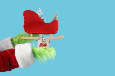 Green hairy hands of creature in costume with Santa sledge and decorative house on blue background clipart