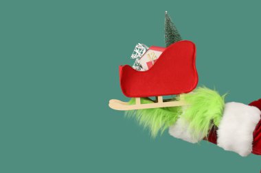 Hairy hands of creature in costume with Santa sledge, decorative fir trees and house on green background clipart