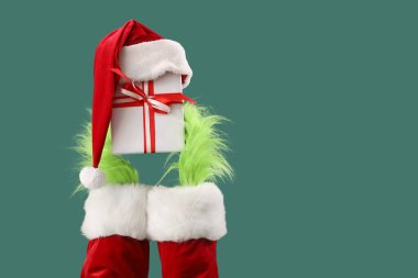 Hairy hands of creature in costume with Santa hat and Christmas gift box on green background clipart