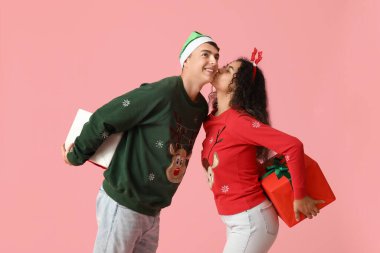 Mutlu genç çift, Noel süveterleri içinde hediye kutularıyla pembe arka planda öpüşüyor.