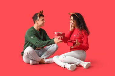 Happy young couple in Christmas sweaters with gift boxes on red background clipart