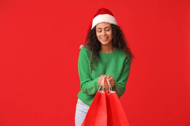 Kırmızı arka planda alışveriş torbaları olan Noel Baba şapkalı güzel bir Afro-Amerikalı kadın. Noel kutlaması
