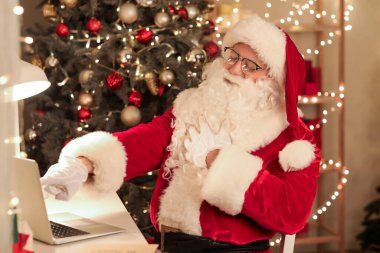 Noel Baba dizüstü bilgisayarla Noel arifesinde evde gülüyor.