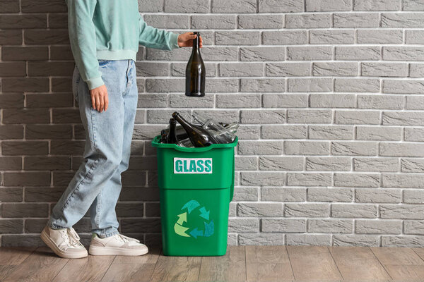 Woman putting bottle into full trash bin with GLASS sign near grey brick wall. Recycling concept