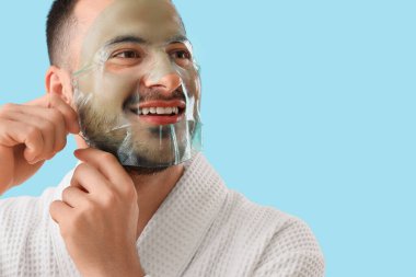 Bornozlu yakışıklı genç adam mavi arka plana kozmetik jel maske takıyor.