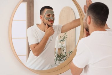 Evdeki banyoda aynanın yanında kozmetik kilden maskeli yakışıklı genç adam.