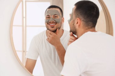 Evdeki banyoda aynanın yanında kozmetik kilden maskeli yakışıklı genç adam.