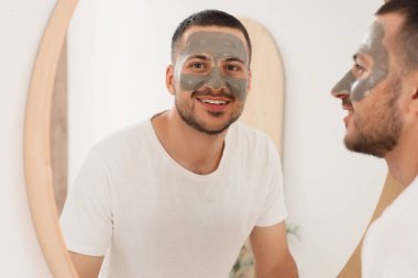 Evdeki banyoda aynanın yanında kozmetik kilden maskeli yakışıklı genç adam.