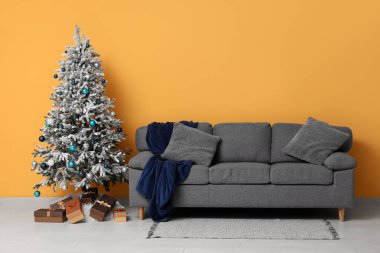 Hediye kutularıyla süslenmiş Noel ağacı, yastıklı kanepe ve sarı duvarın yanında ekose.