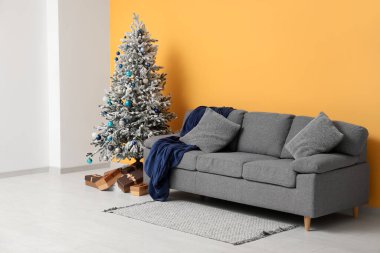 Hediye kutularıyla süslenmiş Noel ağacı, yastıklı kanepe ve sarı duvarın yanında ekose.