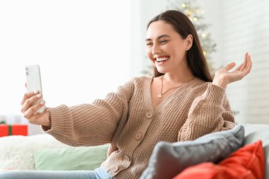 Güzel genç bir kadın selfie çekiyor ve oturma odasında kanepede oturuyor. Noel kutlaması