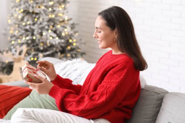 Noel hediyesi kutusuyla oturma odasındaki kanepede oturan genç ve güzel bir kadın.