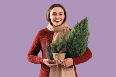 Genç bir kadın, saksıda Noel ağacı ve mor arka planda dalları var.