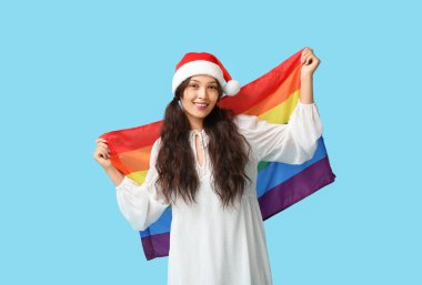 Mavi arka planda LGBT bayrağı olan Noel Baba şapkalı genç Asyalı kadın.