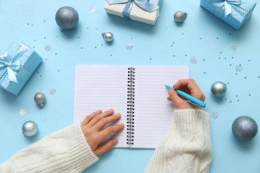 Not defterine 2025 yıllık planlar yazan bir kadın. Noel baloları ve mavi arka planda hediyeler.