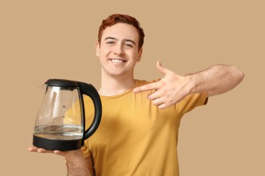 Yakışıklı genç adam bej arka planda modern elektrikli su ısıtıcısını işaret ediyor.