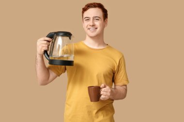 Modern elektrikli su ısıtıcısı ve bej arka planda fincanı olan yakışıklı genç adam.