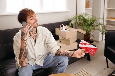 Elinde hediye çeki olan genç dövmeli adam evdeki kanepede cep telefonuyla konuşuyor.