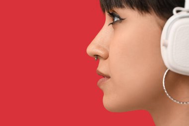 Kırmızı arka planda burun piercingli, kulaklıklı şık genç bir kadın.