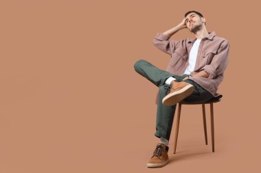 Şık giyinmiş yakışıklı genç adam taburede oturuyor ve bej arka planda poz veriyor.