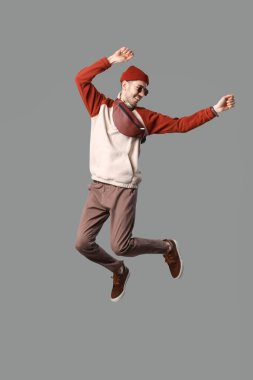 Yakışıklı, güler yüzlü, şık elbiseli, güneş gözlüklü ve çantalı, gri arka planda atlayan genç adam.