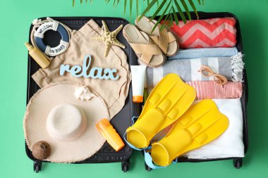 Yeşil arka planda plaj aksesuarları, kıyafetler ve kozmetik ürünlerle dolu bavulu aç