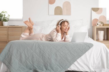 Kulaklığı ve laptopu olan genç bir kadın yatak odasında yatıyor.
