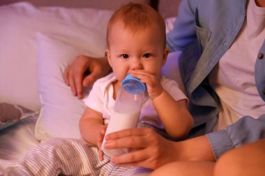 Anne, gece yatak odasında küçük bebeğini biberonla besliyor.